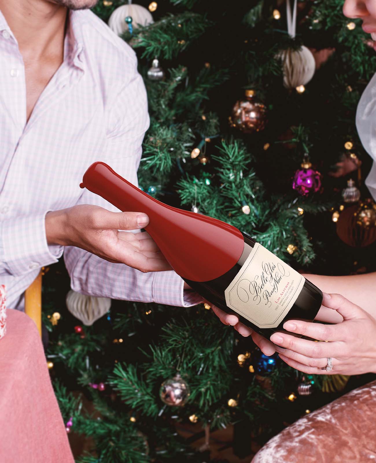 Two people exchanging a wax-dipped wine bottle in front of a decorated Christmas tree, holiday gifting moment.