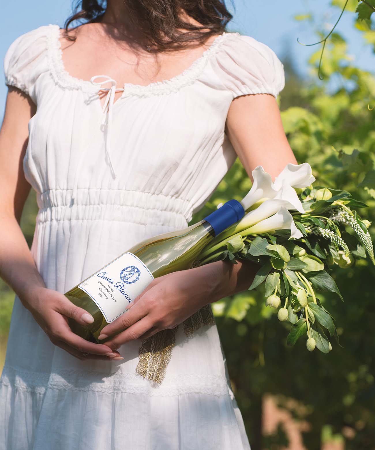woman in a white dress in a vineyard holding a bouquet of flowers and a bottle of Chardonnay wine