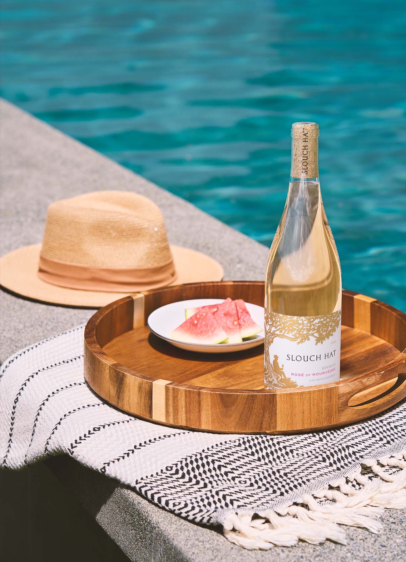 Slouch Hat Rosé of Mourvèdre wine bottle in an outdoor poolside lifestyle scene on a wooden tray with a straw hat and watermelon, created by Outshinery
