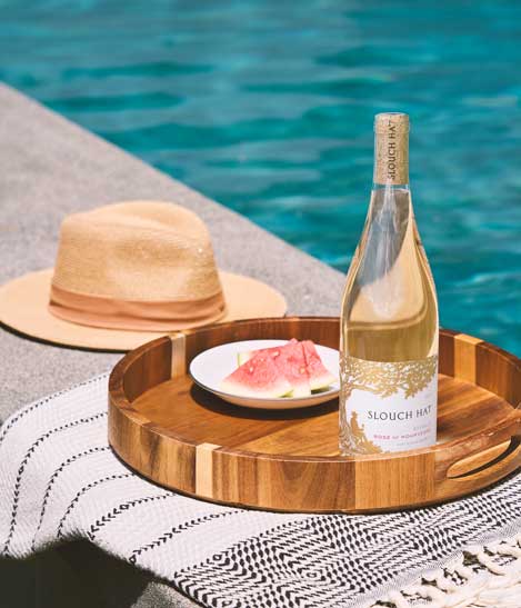 Slouch Hat Reveille Rose of Mourvedre bottle on a poolside tray with watermelon slices and a straw hat, by Outshinery