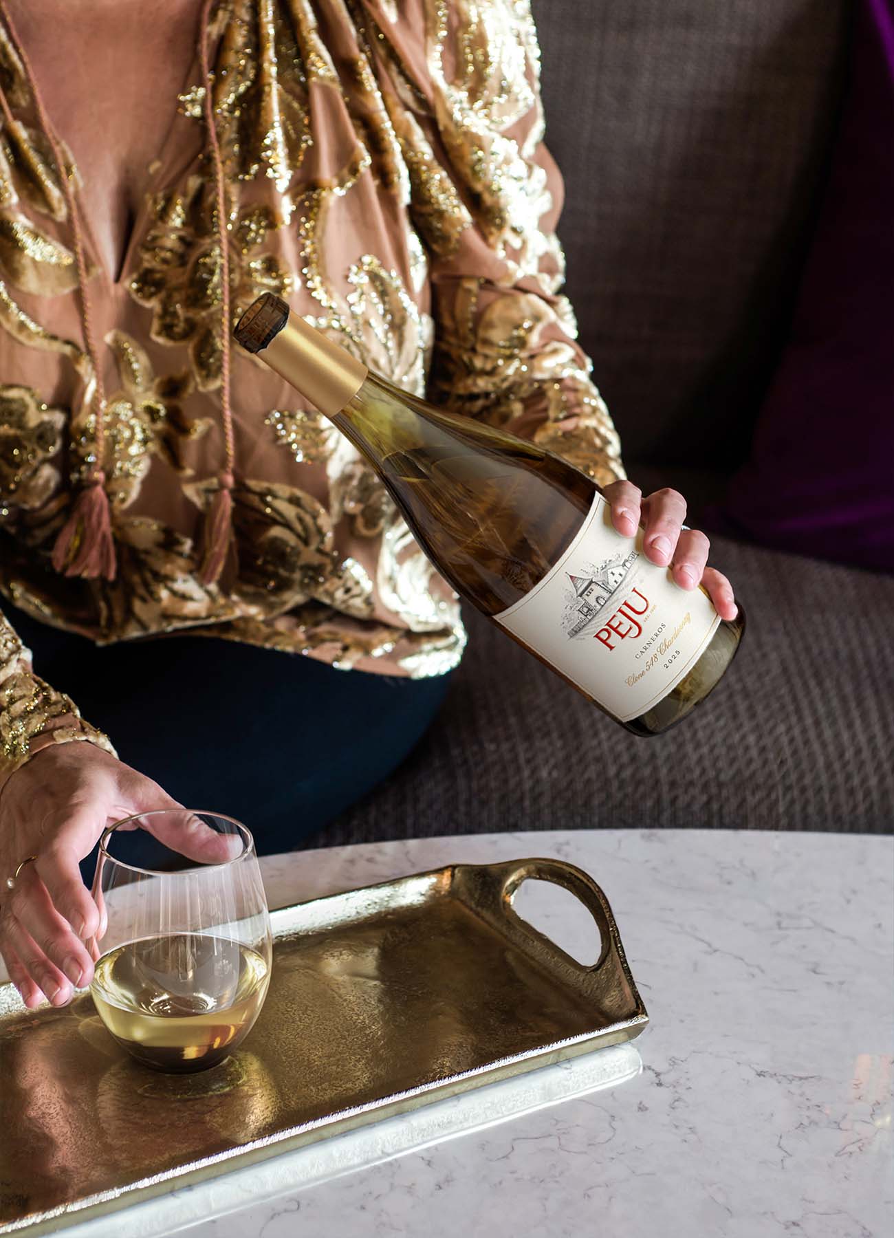 A woman in a gold sequined top pours Peju Carneros Chardonnay into a stemless glass on a marble table, styled for a holiday gathering