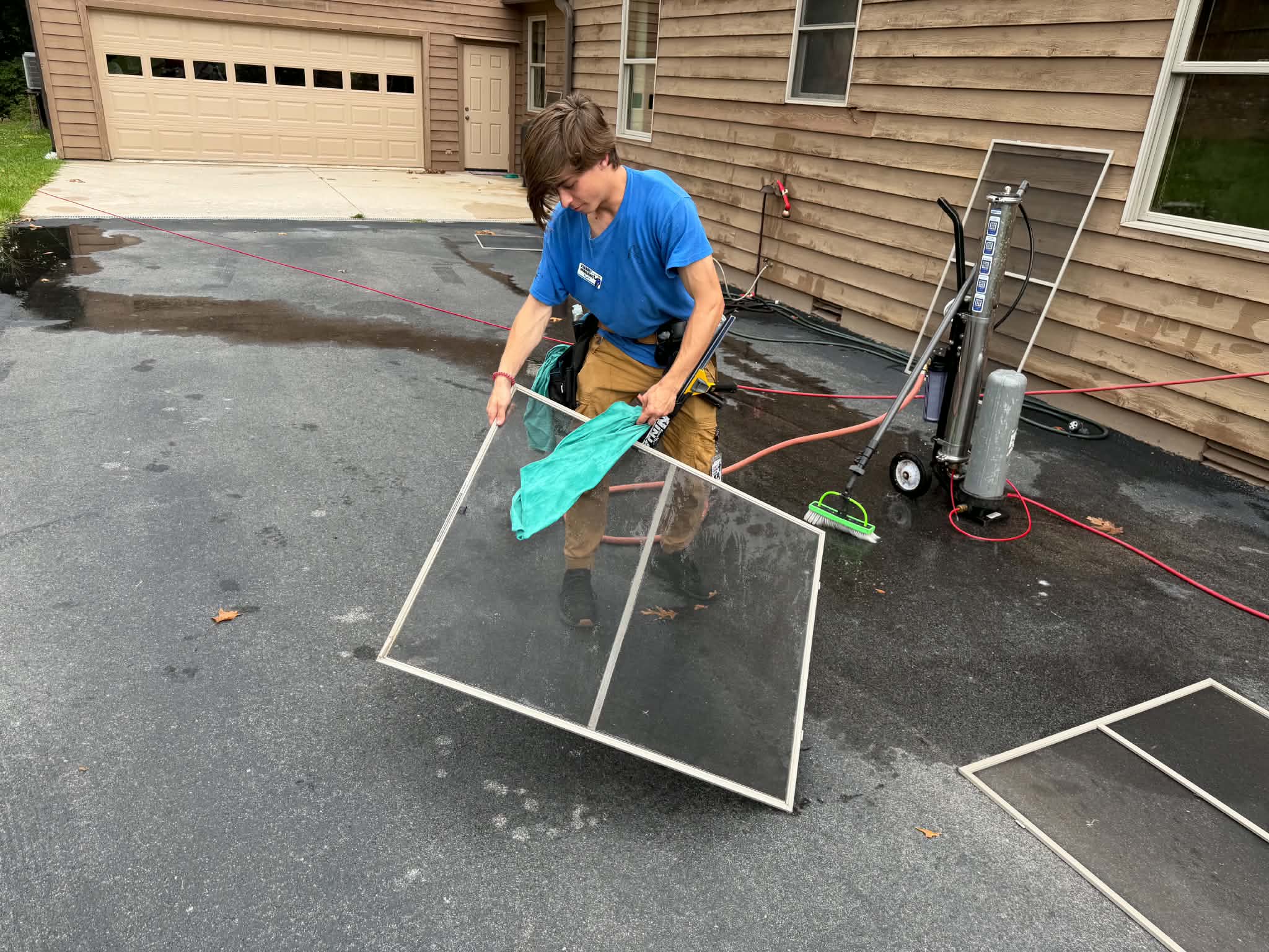 Man wearing Clearly Windows uniform cleaning a window screen outside a house with cleaning supplies nearby.