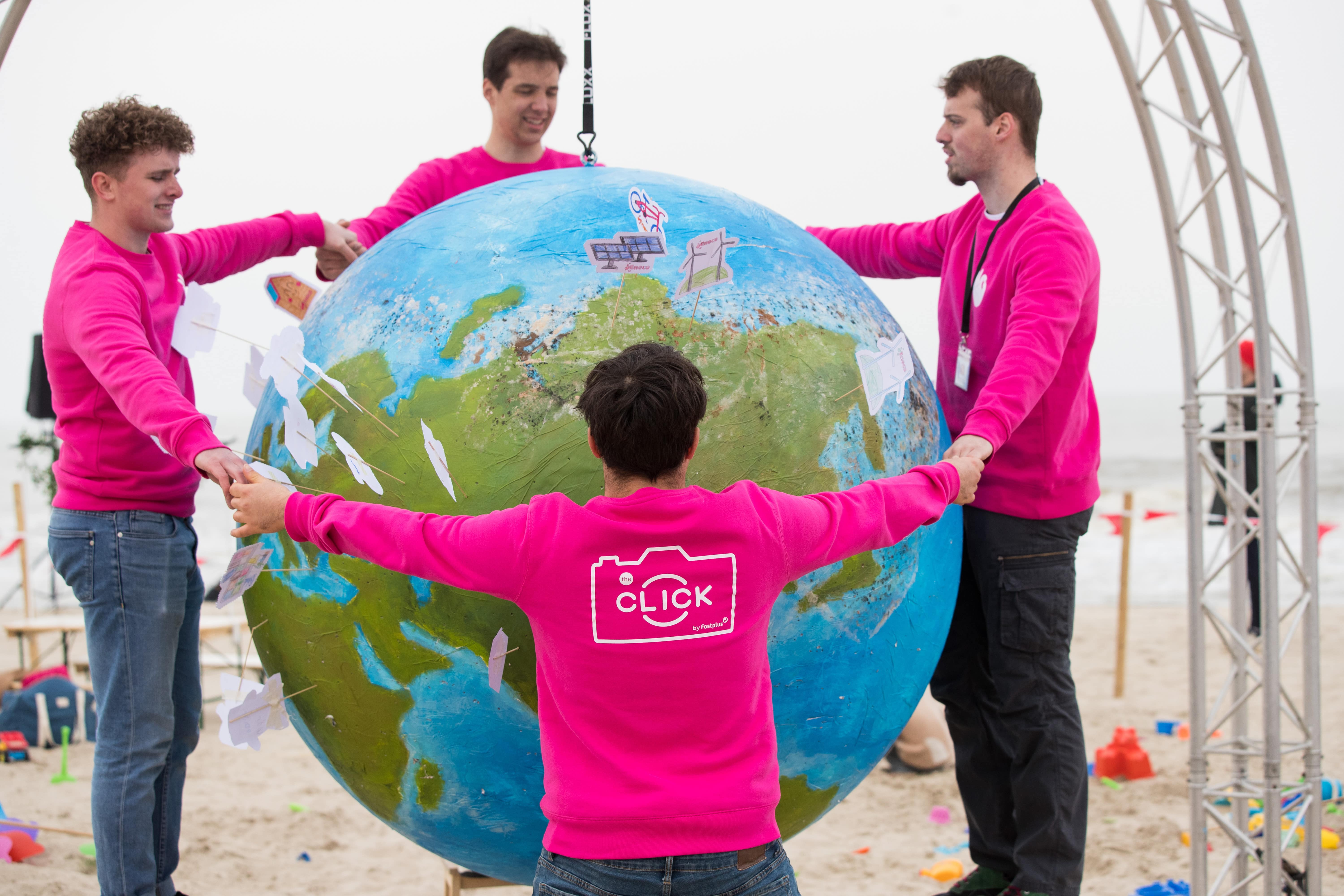 Field & Concept brand ambassadors in pink 'The Click by Fost Plus' sweatshirts holding a large painted Earth globe during a beach recycling awareness activation