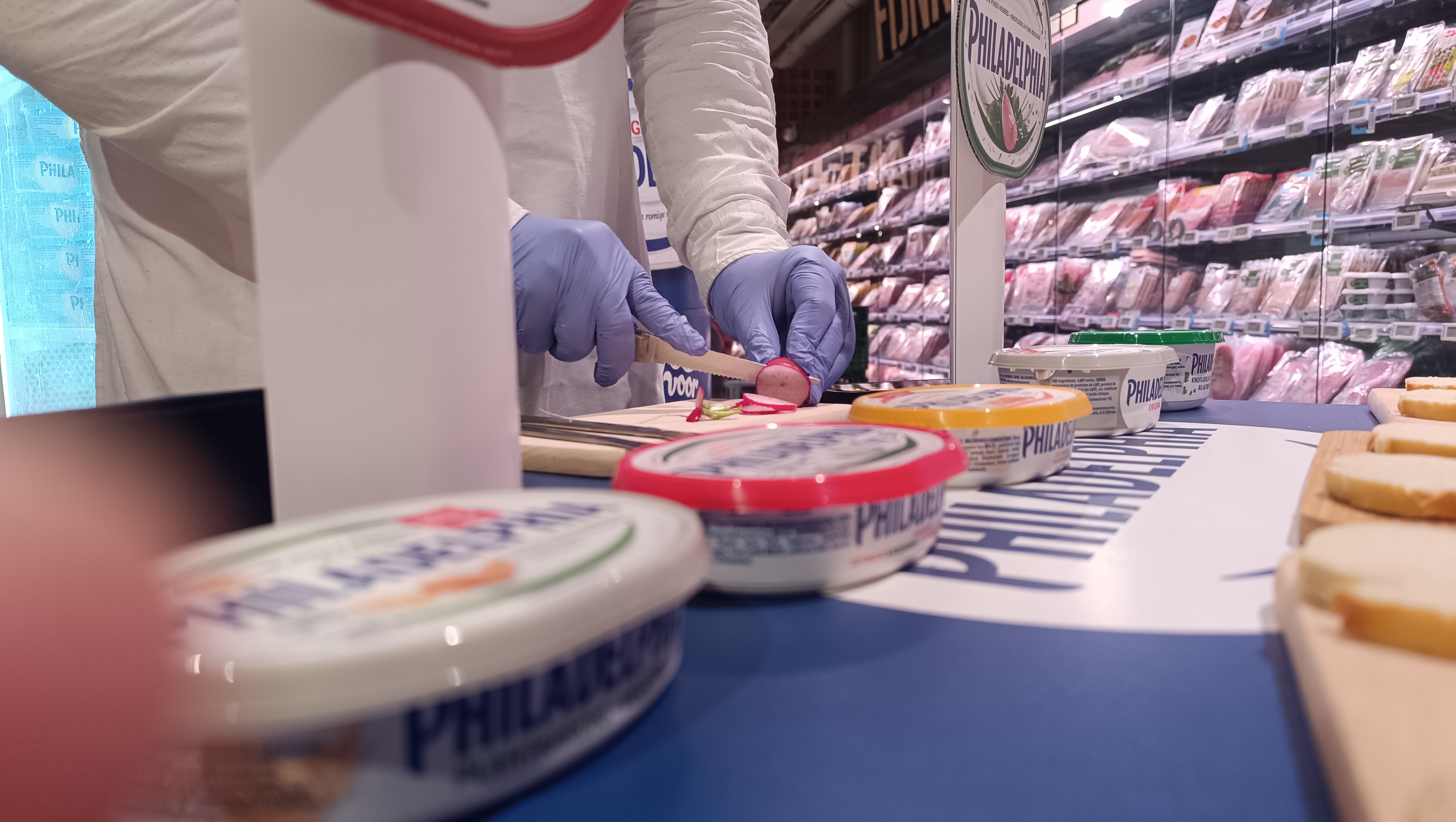 Close-up of a brand ambassador slicing radish for a Philadelphia cream cheese tasting demo, with sample tubs and bread slices laid out on the counter