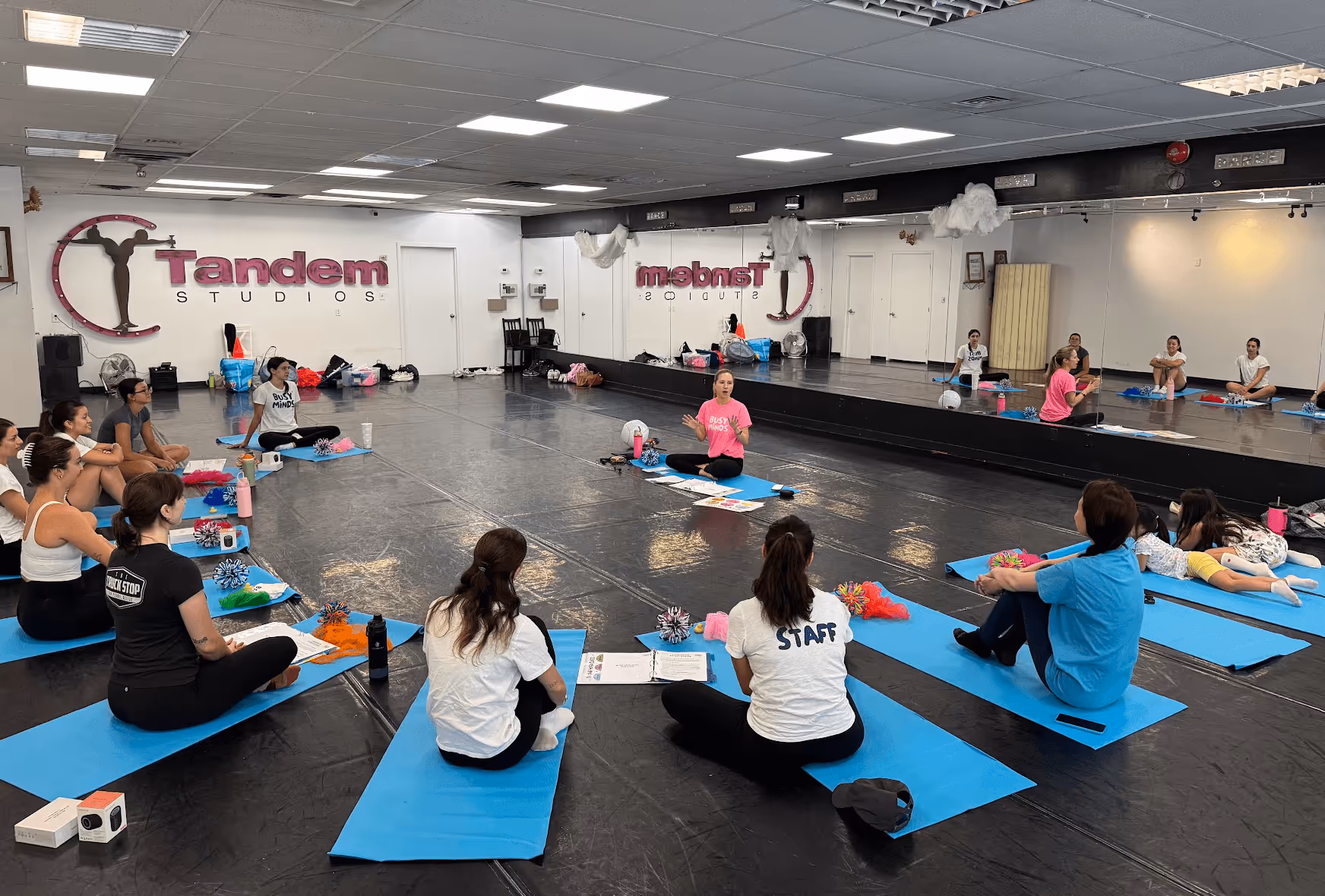 A group of people learning how to operate a Busy Minds Franchise. It is a class, people are sitting on yoga mats in a circle listening to a person standing and speaking in the middle.