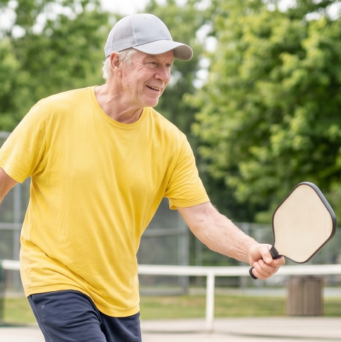 Older man wearing yellow shirt and gray cap playing pickleball outdoors with paddle in hand.