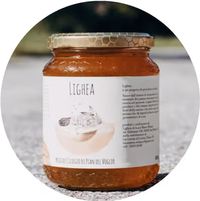 A glass jar of Lighea honey with a gold lid. The label features a light-colored background and text describing the product. The jar is placed on a stone surface, surrounded by a natural setting.
