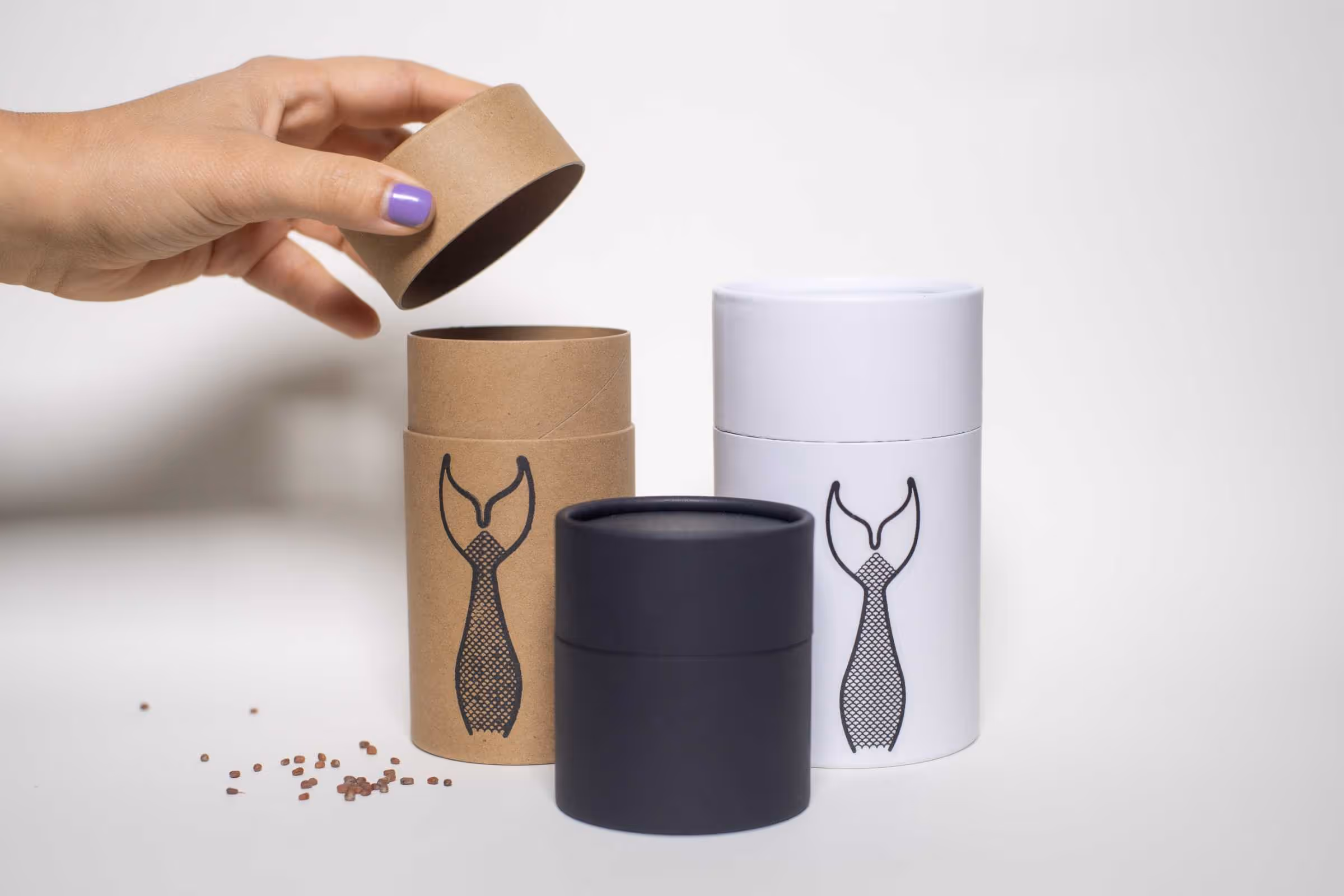 A hand lifting the lid off a brown cylindrical container. Next to it are two additional containers, one white and one black, each featuring a graphic of a tie. Small particles are scattered on the surface below.