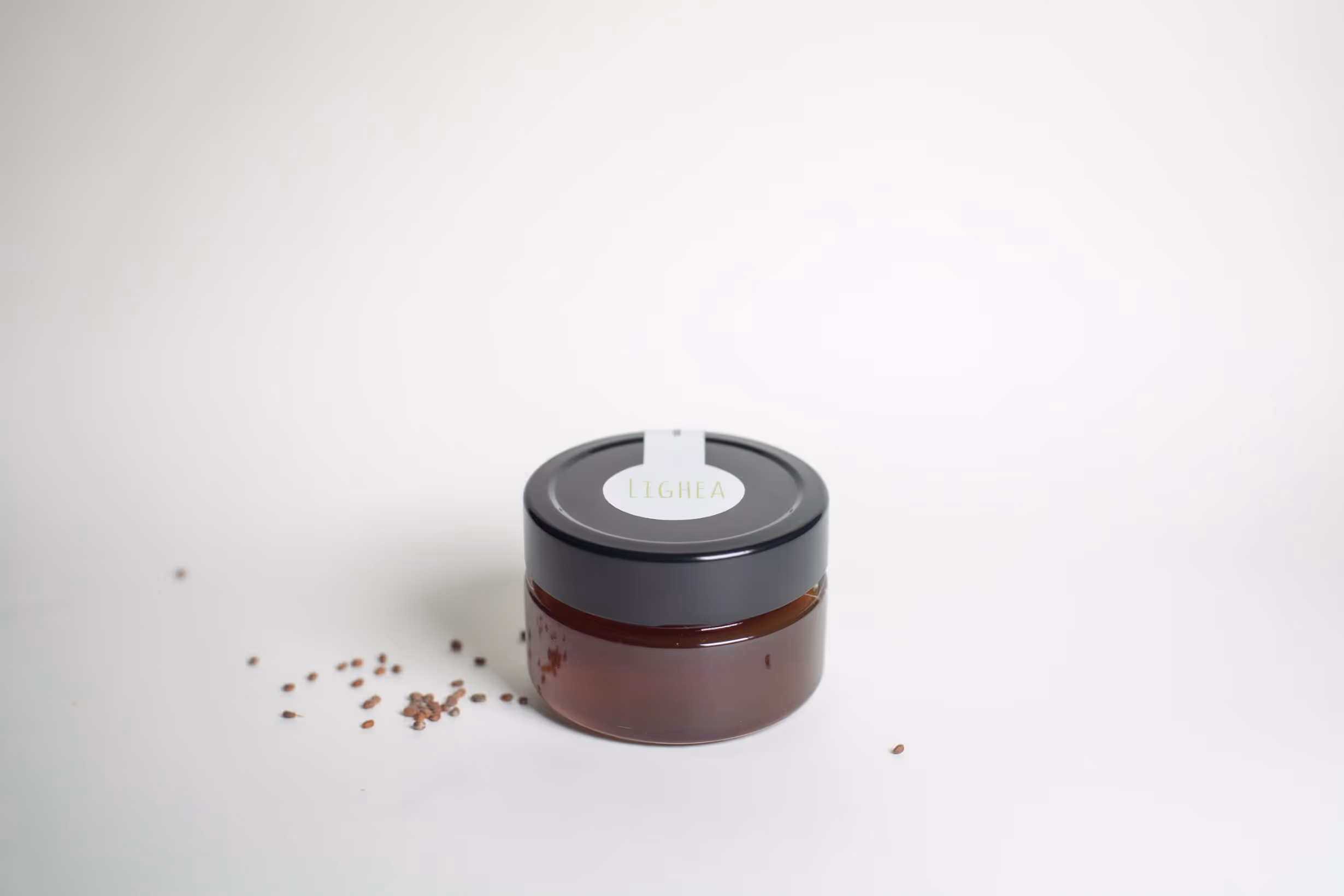 A small amber glass jar with a black lid and a white label, placed on a white background. Some small seeds are scattered around the jar.