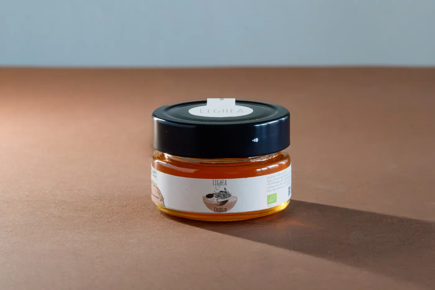 A small amber glass jar with a black lid and a white label, placed on a white background. Some small seeds are scattered around the jar.
