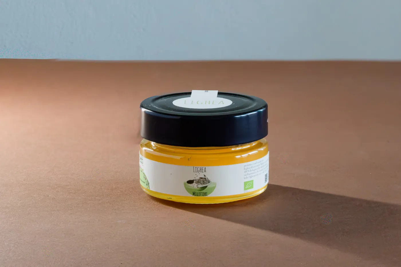 A glass jar of Lighea honey placed on a wooden surface, featuring a colorful label with illustrations and text describing the product. The jar has a metallic lid, and the background is softly blurred.
