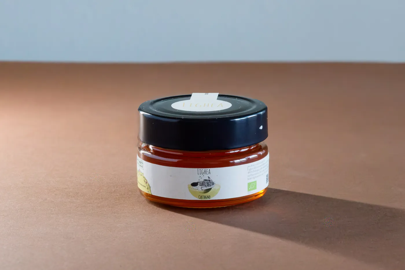 A glass jar of Lighea honey with a gold lid. The label features a light-colored background and text describing the product. The jar is placed on a stone surface, surrounded by a natural setting.