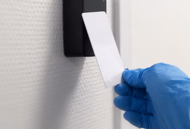 Hand wearing a blue glove holding a white access card near a wall-mounted card reader.