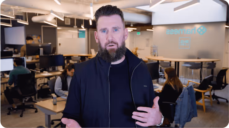 Bearded man speaking in modern open office with people working at desks in the background.