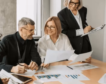 Three professionals reviewing and discussing documents with charts and graphs around a wooden table in a bright office.