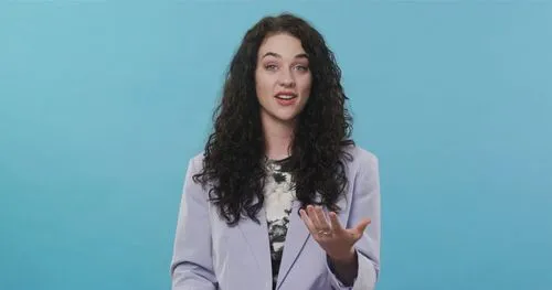 A woman speaking on camera in front of a solid blue background, presenting directly to viewers.