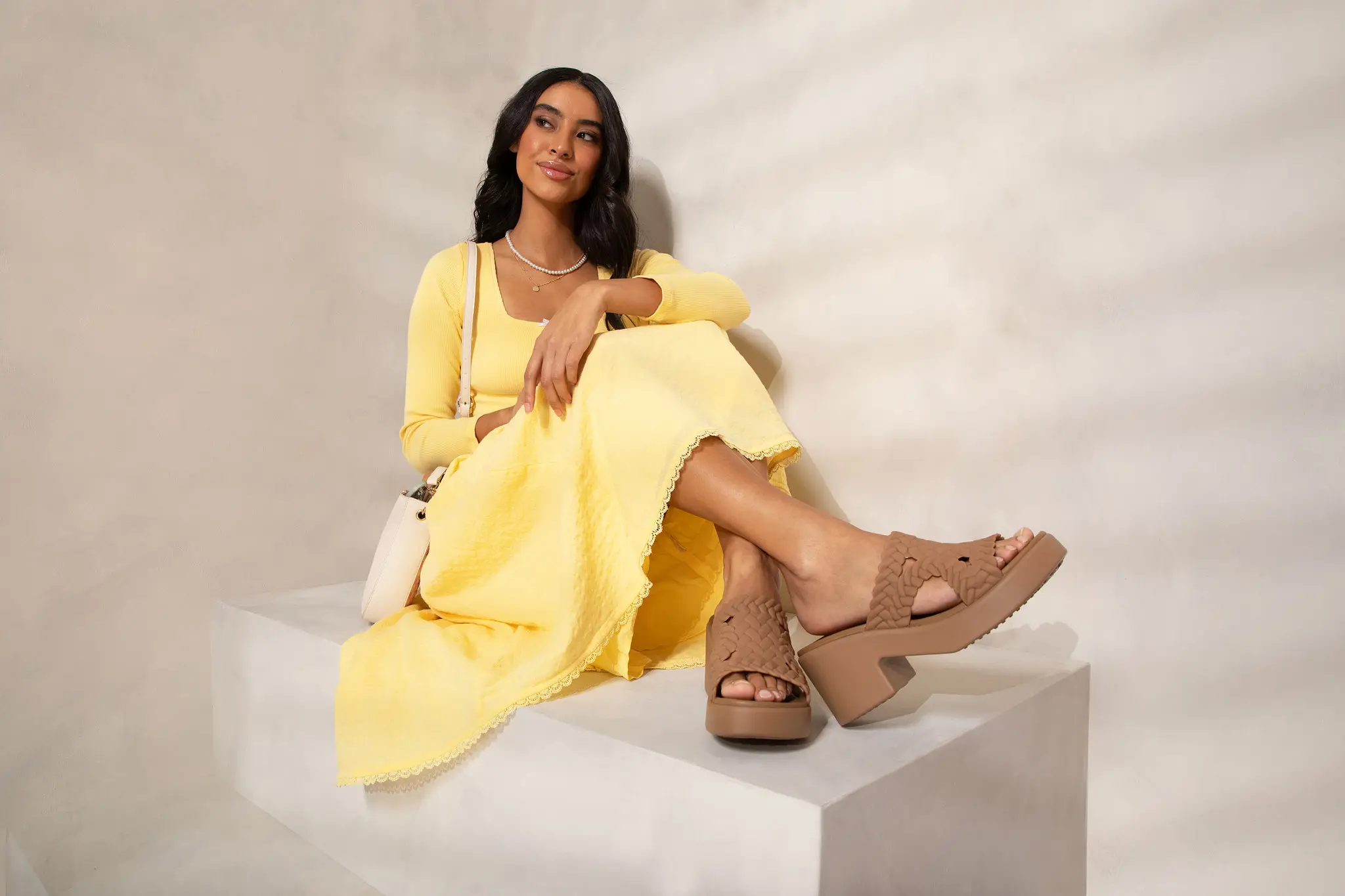 A female model sitting on a step wearing a yellow sundress and Crocs with heels.