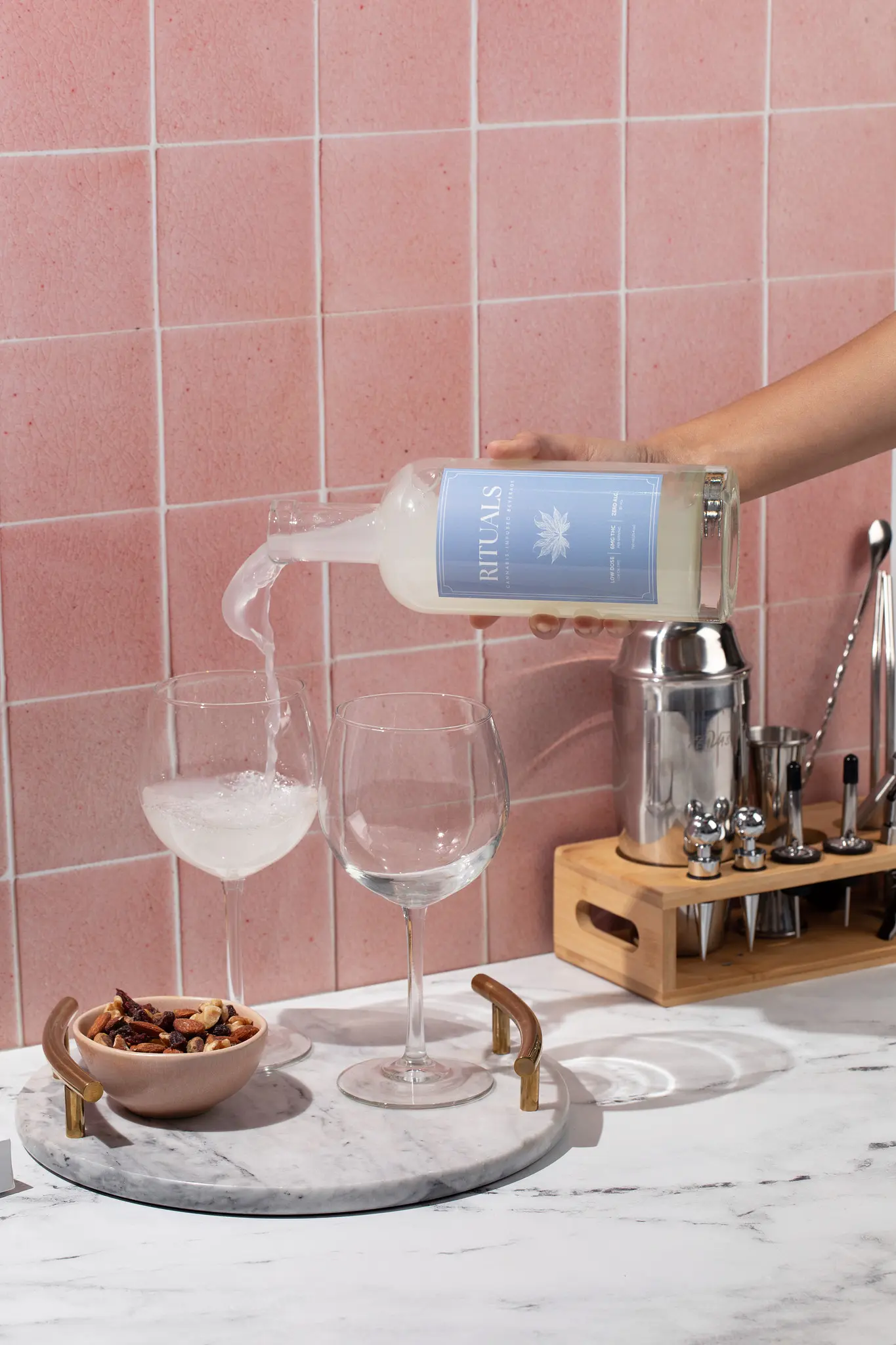 a hand pouring a bottle of liquid into a wine glass with a pink tile backdrop