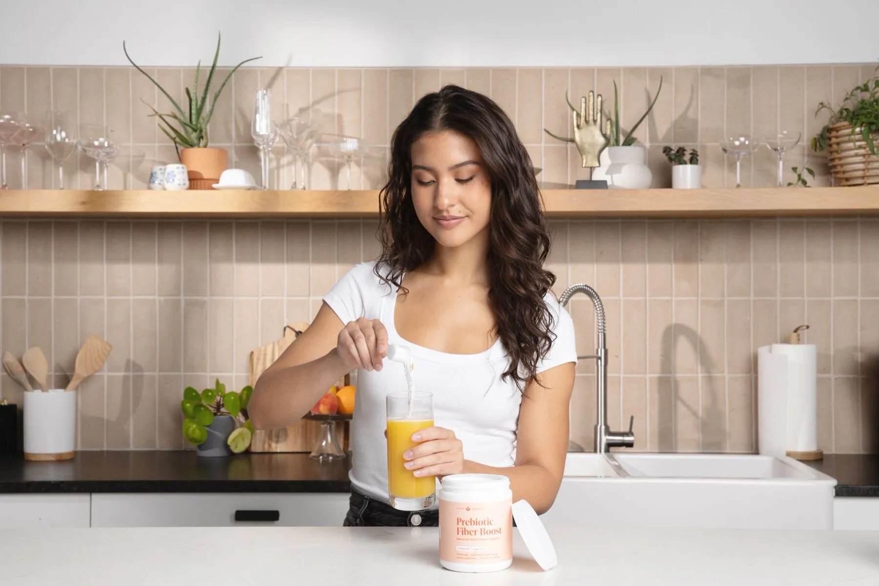 woman pouring prebiotic fiber boost powder into a glass of orange juice in a pretty kitchen