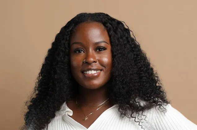 Smiling Black woman with long curly hair wearing a white sweater against a beige background.