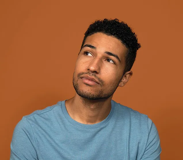 Young man with curly hair and light beard wearing a blue shirt, looking thoughtfully upward against a solid brown background.