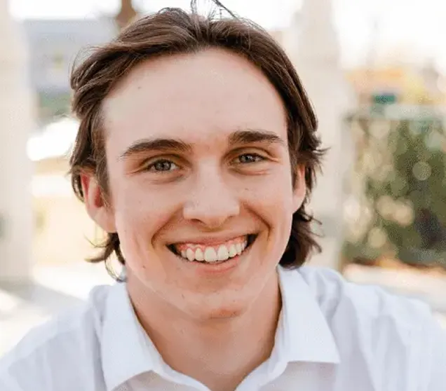 Young man with medium-length brown hair smiling and wearing a white shirt with a blurred outdoor background.