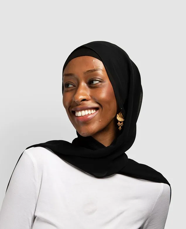 Smiling woman wearing a black hijab and gold earrings, looking off to the side against a light gray background.