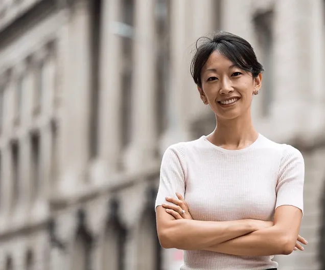 Smiling woman with short black hair, wearing a light pink short-sleeve top, standing with arms crossed in front of a blurred classical stone building.