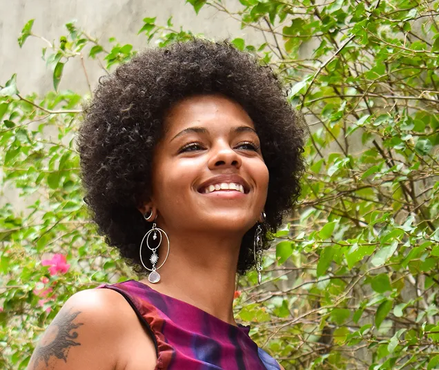 Smiling woman with curly afro hair wearing large silver earrings and a colorful sleeveless top, standing outdoors with green foliage in the background.