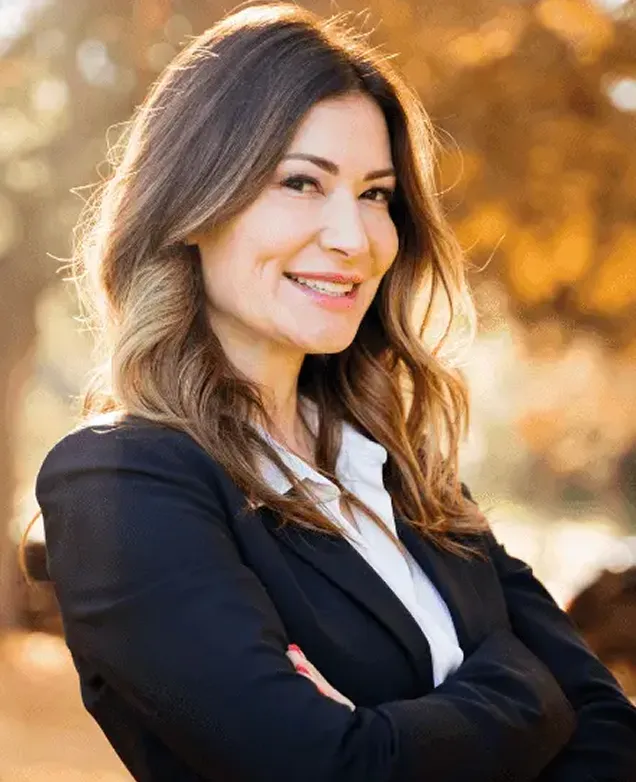 Smiling woman with braces wearing a black blazer and white shirt, standing outdoors with arms crossed and autumn foliage in the background.