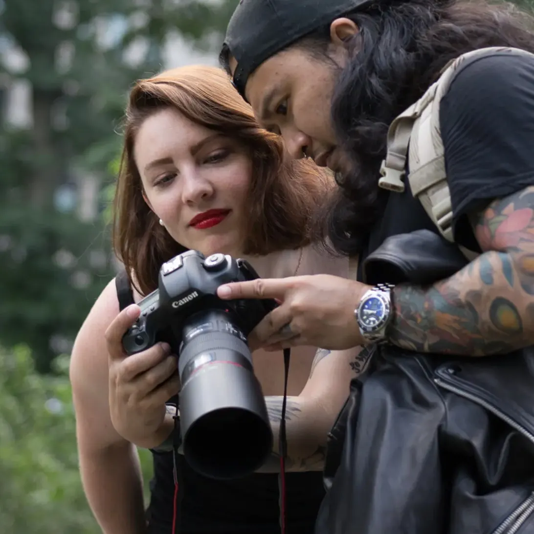 Two photographers, a woman and a man with tattoos, reviewing photos on a Canon camera outdoors.