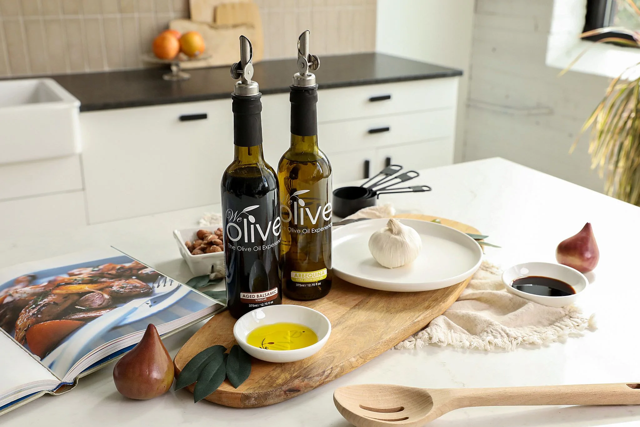 Kitchen countertop with olive oil bottles, garlic, figs, measuring cups, cookbook, and wooden spoon.