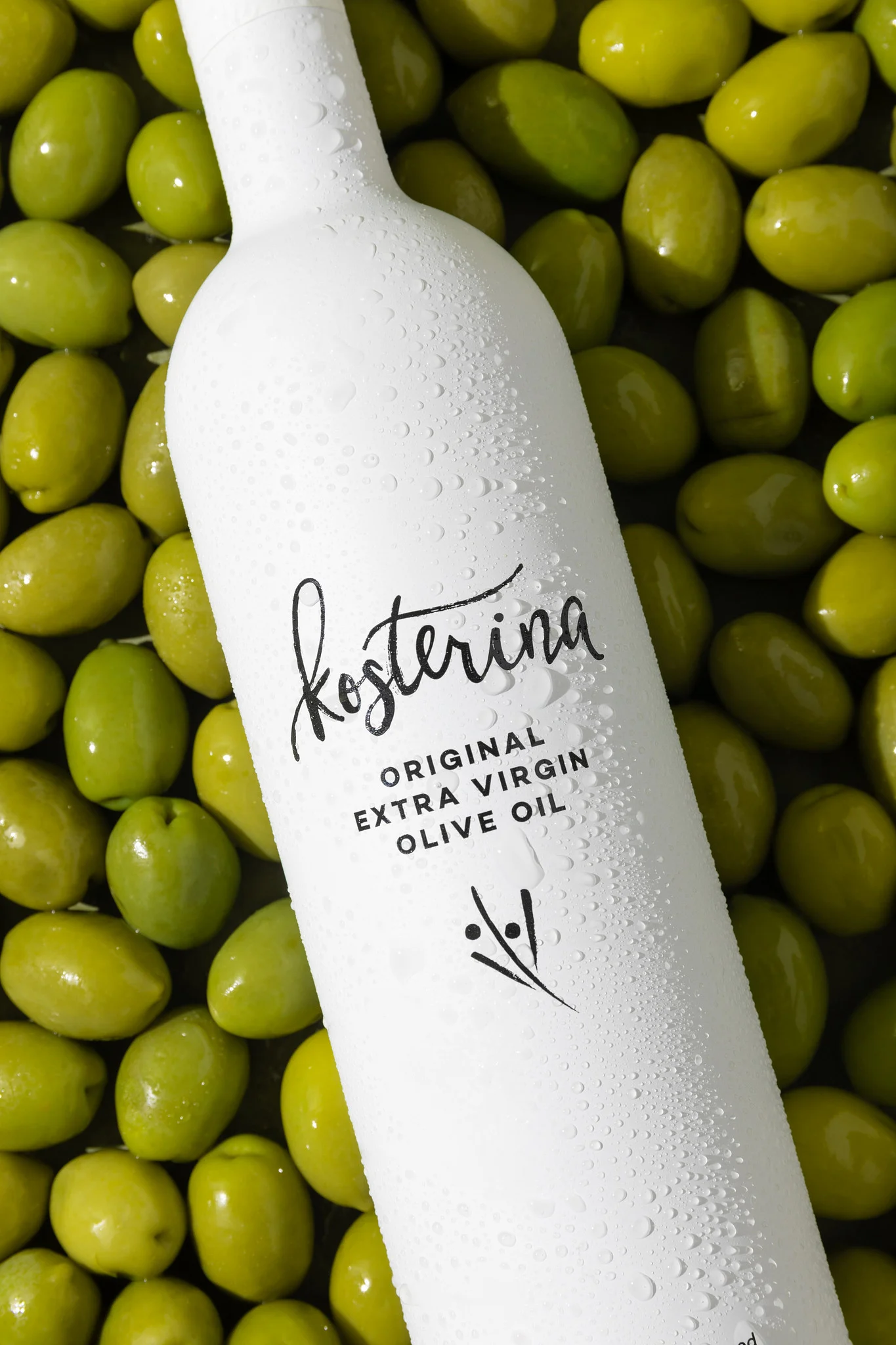 White bottle of Kosterina Original Extra Virgin Olive Oil on green olives with water droplets.