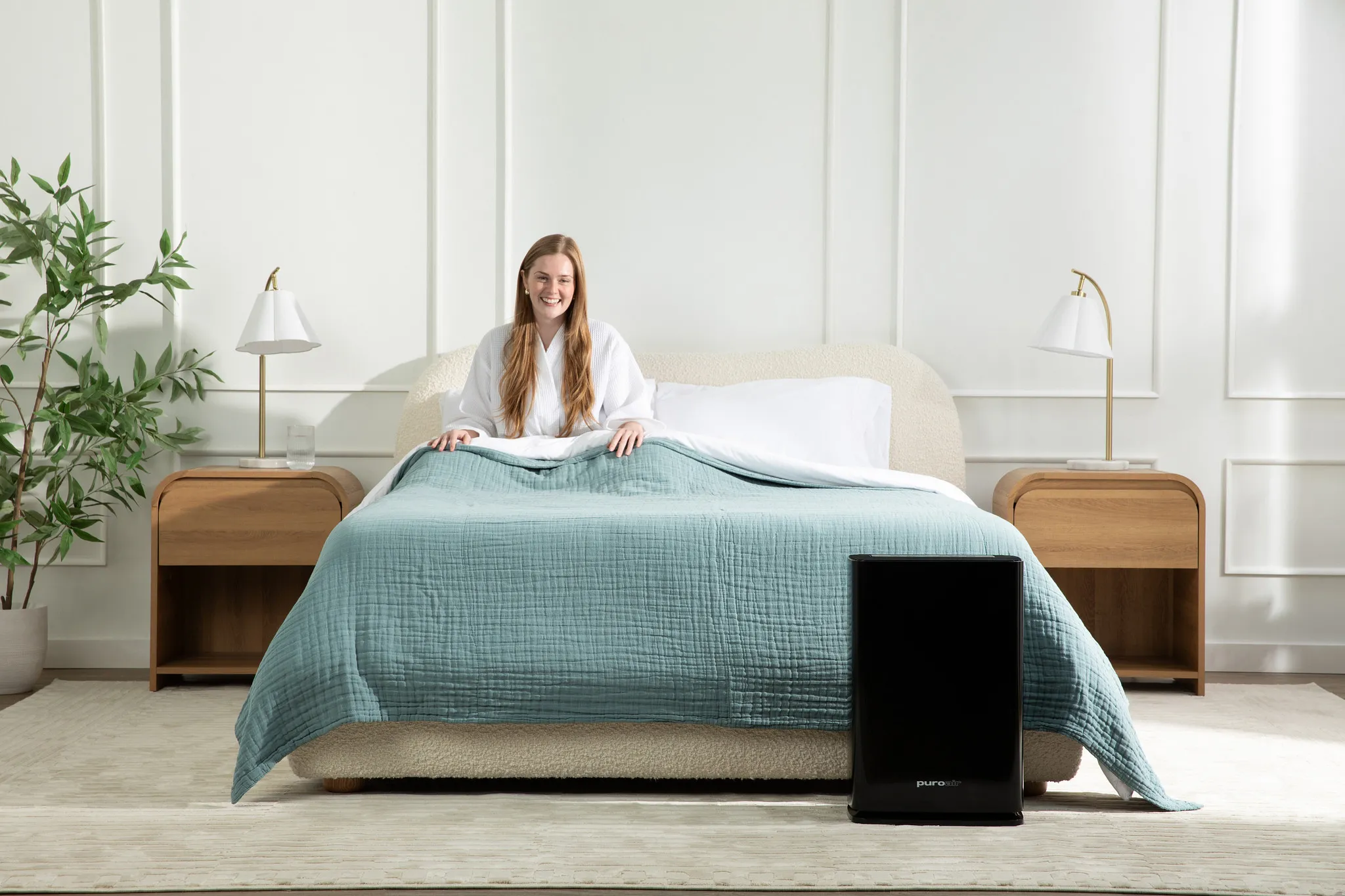 Smiling woman in white robe sitting in bed with teal quilt and air purifier on floor in a bright room.