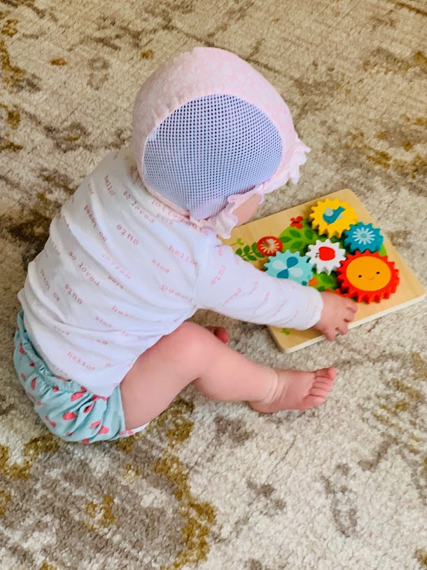 Baby wearing hearing aids and a pilot cap sitting on floor playing with wood toy gears