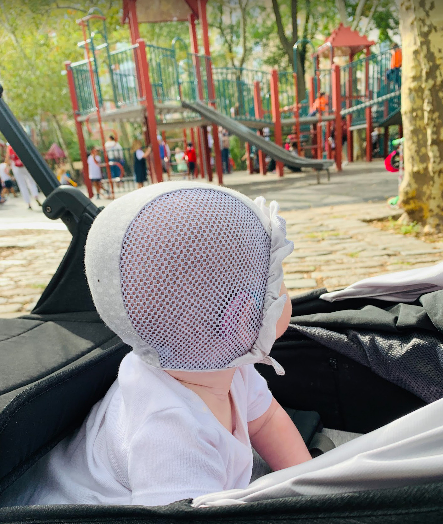 Baby wearing hearing aids and pilot cap sitting in a stroller looking at a playground scene