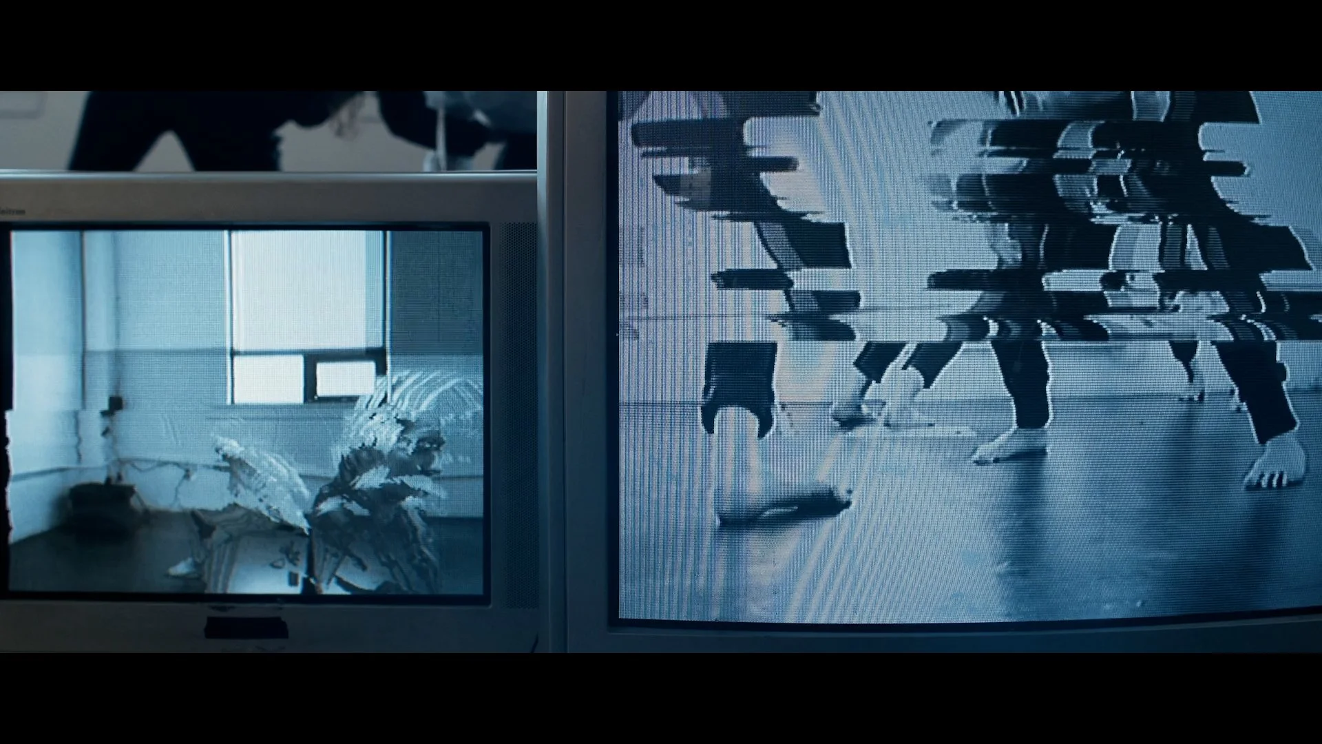 Two CRTs side by side showing two blue-grey images. The right one shows a glitched-out shot of a few people dancing from the waist down. The other depicts a sparse room with a window in the background with two glitchy and distorted figures in the foreground. 