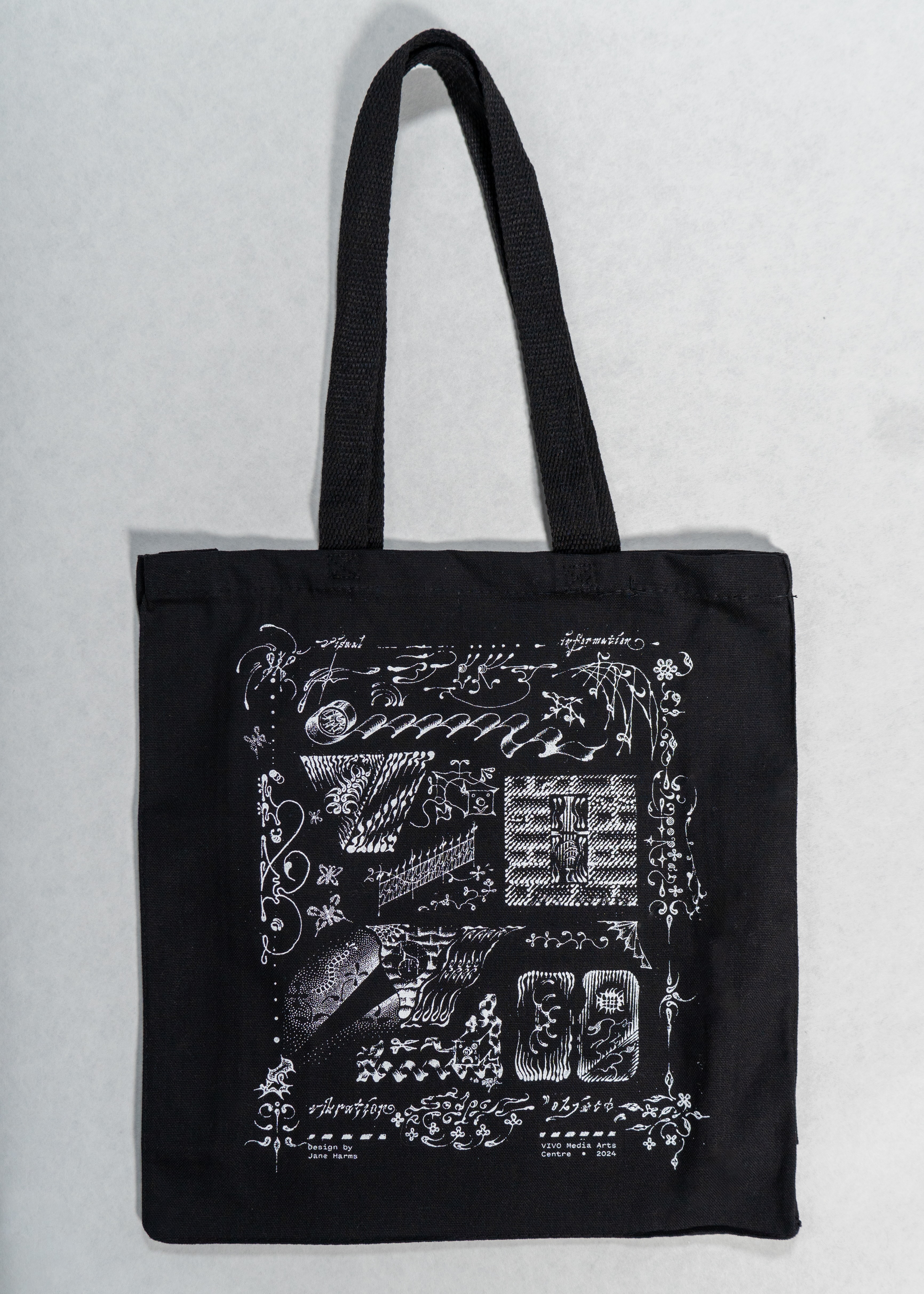 A black canvas tote bag with a white print featuring a variety of stylized sketch-like abstract drawings, including vines, music notes, letters and flowers.