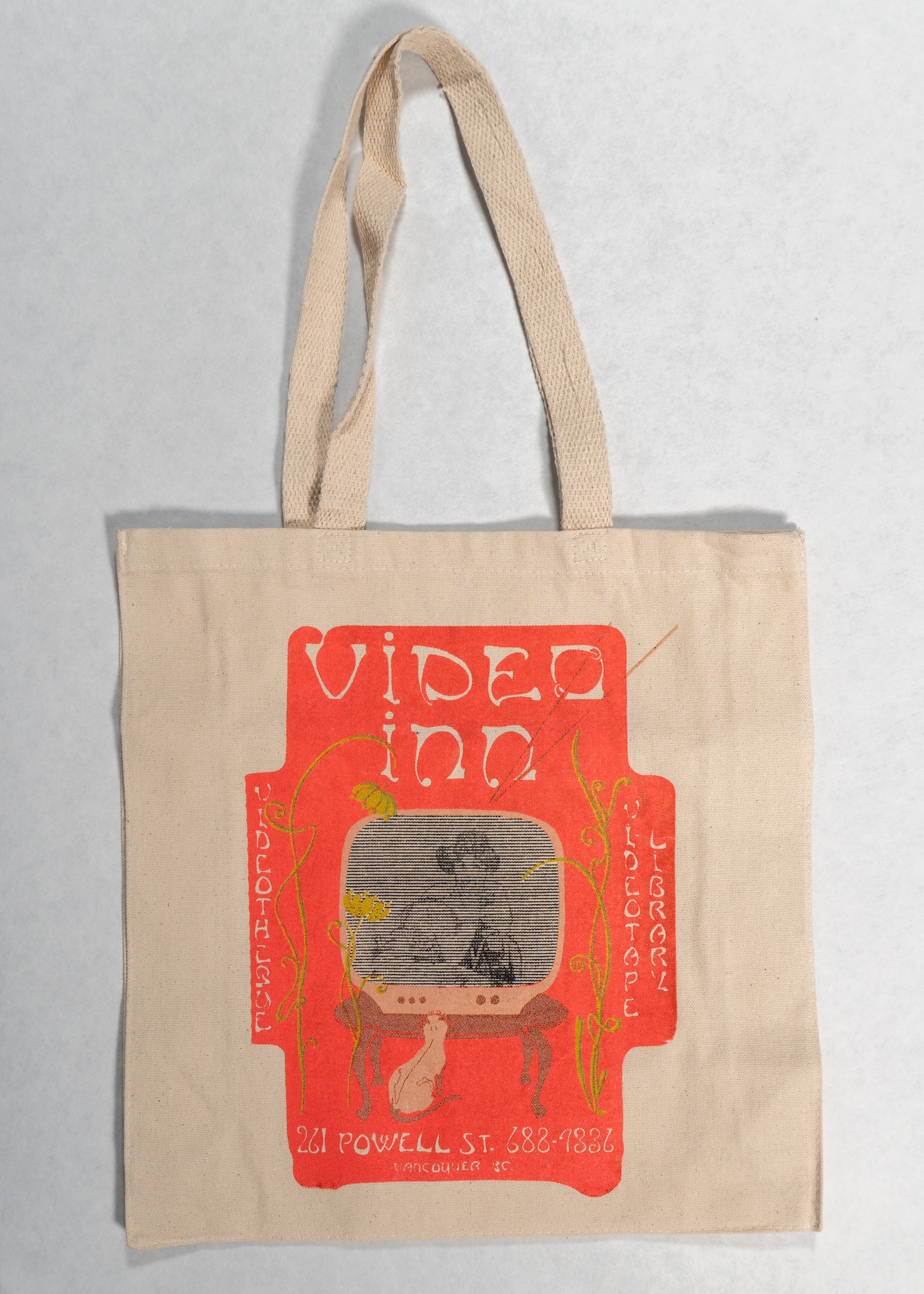A beige cloth tote on a white background with a pink rectangular print on it. The print reads “ViDEO iNN” at the top in flowery text, and there is a cat watching a retro tv set surrounded by three flowers with long stems. 