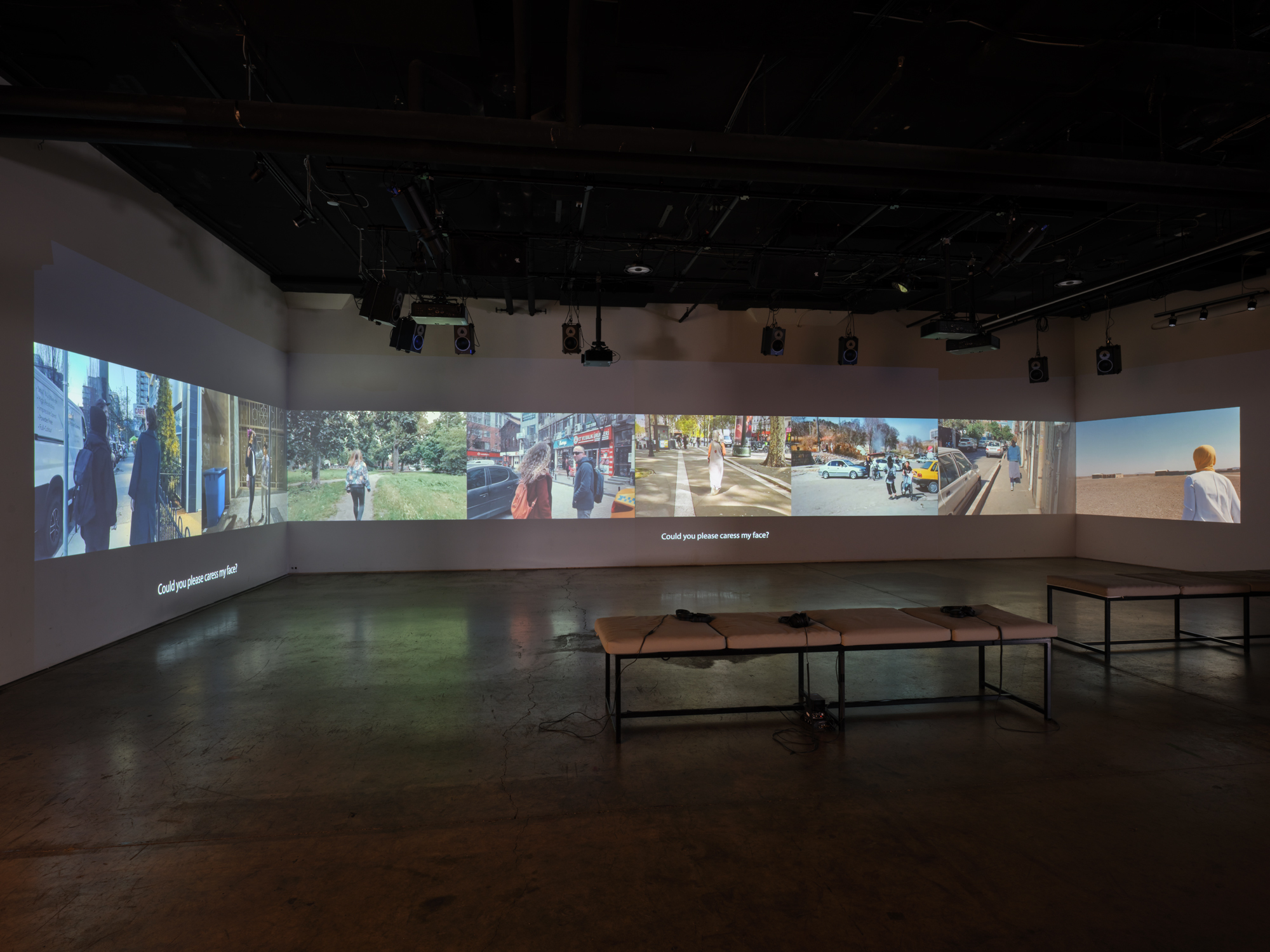 A completely dark studio room with projections on all three visible walls. On the projections are multiple different images of people in everyday scenarios, including walking in the park or on the street. In the room are two cushioned benches positioned near the middle of the space. 