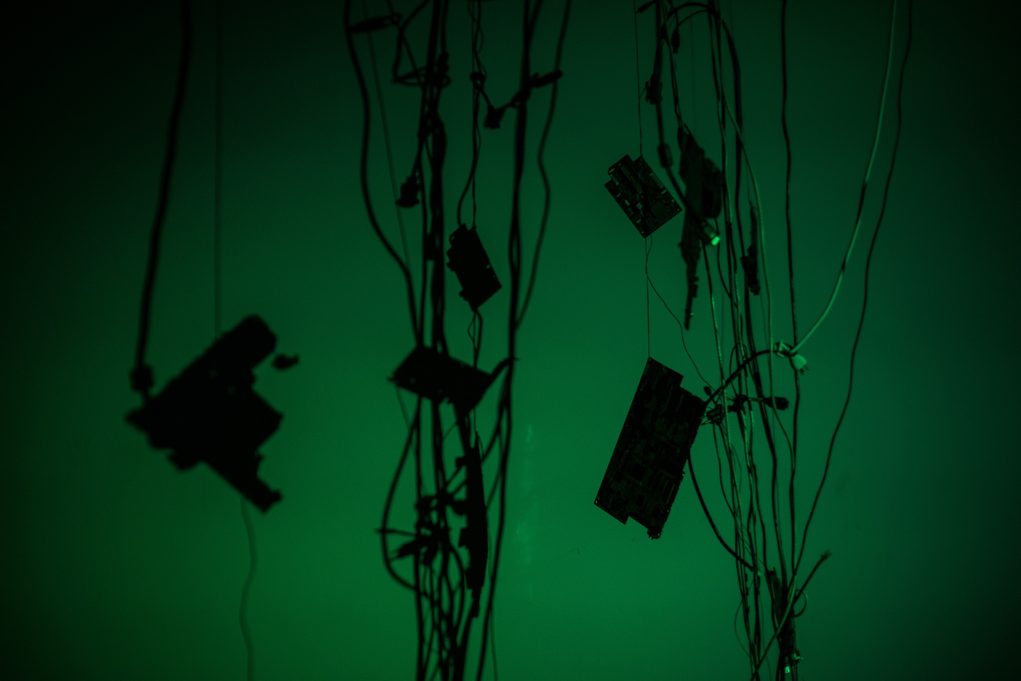Multiple disassembled components of technological devices attached to cords suspended in a room dimly lit by an ominous green light. 