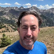 Man with short dark hair and beard wearing a blue shirt smiling outdoors with mountain peaks and a partly cloudy sky in the background.
