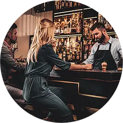 Bartender talking to a woman seated at a bar counter with a man sitting next to her, shelves with bottles in the background.