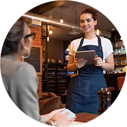 Smiling waitress in denim apron holding a tablet while taking an order from a seated customer in a restaurant.