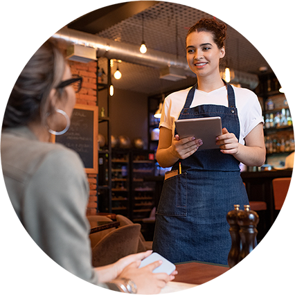 Smiling waitress in denim apron holding a tablet while taking an order from a seated customer in a restaurant.