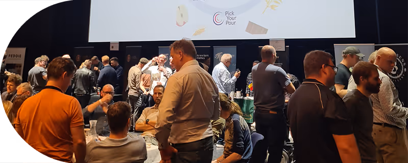 Group of people mingling and tasting drinks at a whiskey festival event with banners and a screen showing 'Pick Your Pour'.