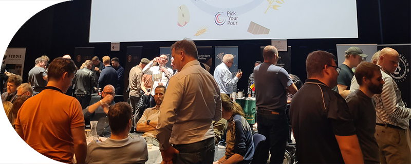 Group of people mingling and tasting drinks at a whiskey festival event with banners and a screen showing 'Pick Your Pour'.
