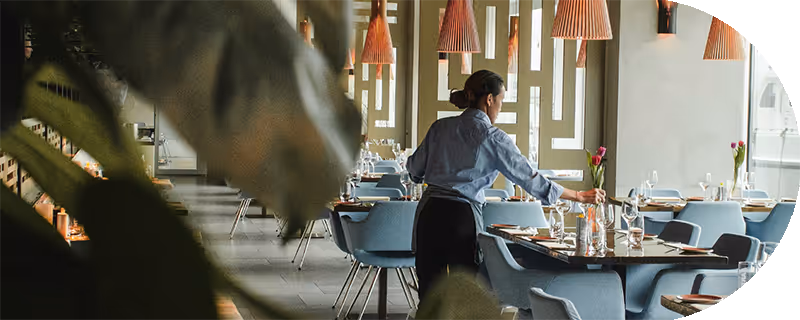 Waitress setting a table in a modern restaurant with blue chairs and pendant lighting.