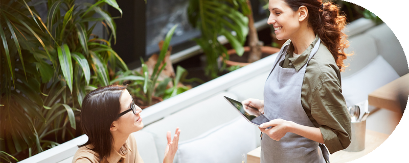 Smiling waitress holding a tablet takes an order from a seated woman wearing glasses in a restaurant with plants in the background.