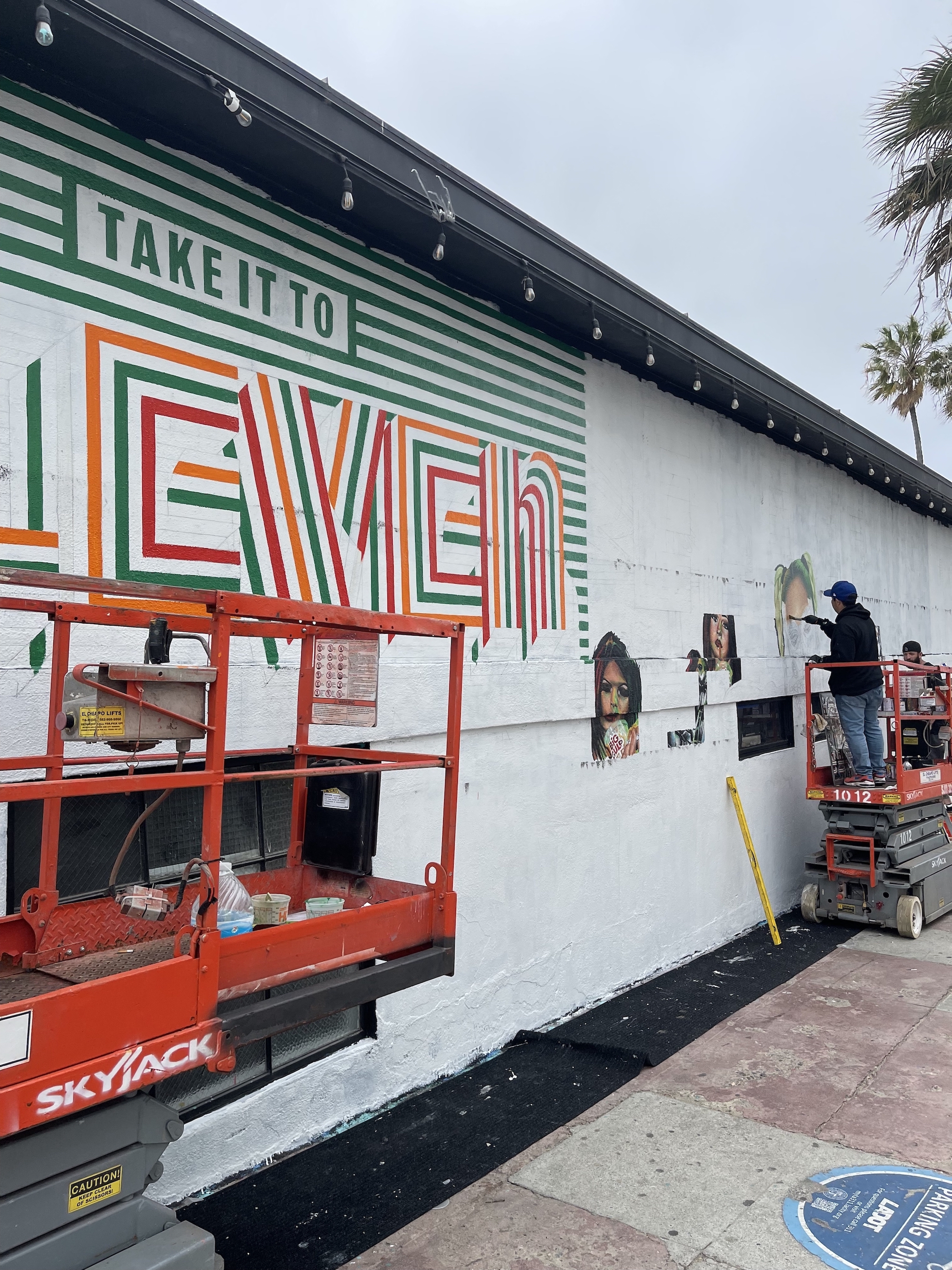 Hoist Media artist on scissor lift painting 7-Eleven mural photoreal portrait on building exterior in Venice CA